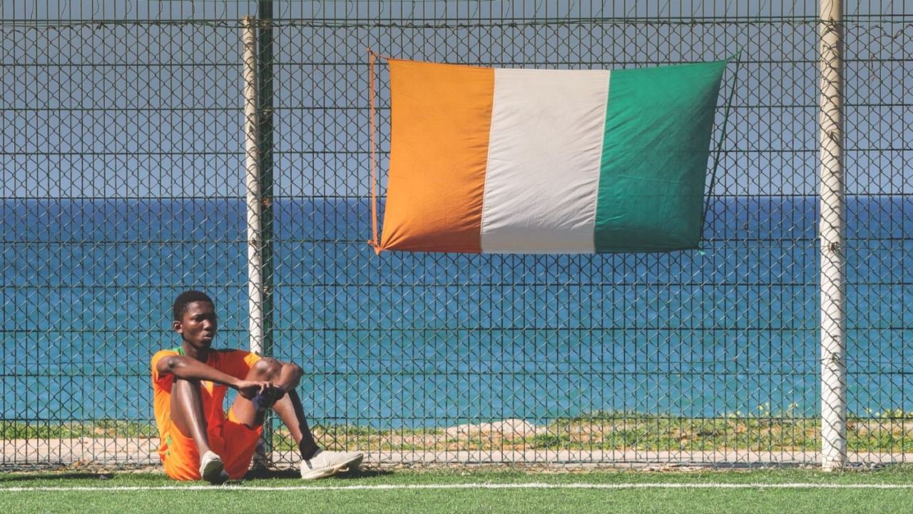 Junger Fußballspieler auf einem Platz am Meer, dahinter die Flagge der Elfenbeinküste