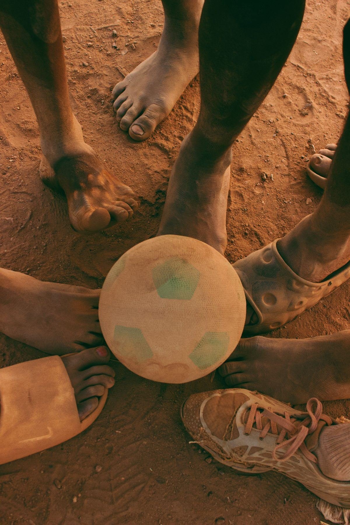 Barfüße und Sandalen im Kreis um einen abgenutzten Fußball auf rotem Sand
