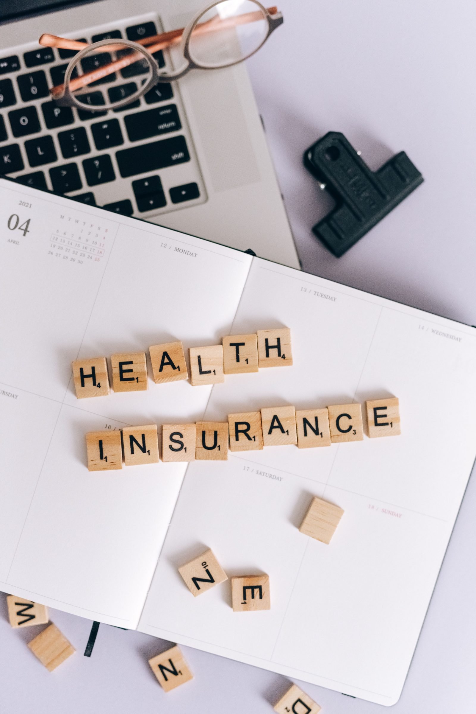 Holzbuchstaben bilden die Worte Health Insurance auf einem aufgeschlagenen Kalender neben Laptop und Brille